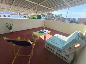 Un balcón techado con una cama, una silla, una mesa y plantas en macetas.