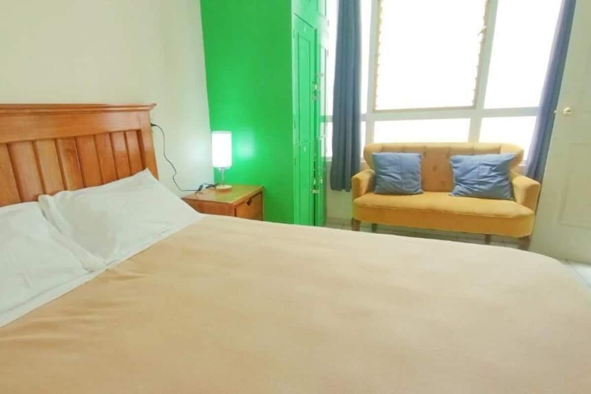 Una cama con cabecera de madera, una mesita de noche con lámpara, pared verde y un sofá amarillo con cojines azules.