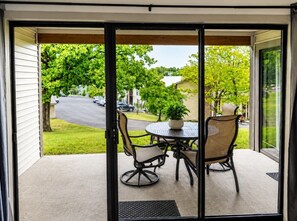 A sliding glass door leading to an outdoor patio with a round table and two chairs, a potted plant, and a view of a street and greenery. - Clubhouse Cove at Pointe Royale (Branson)