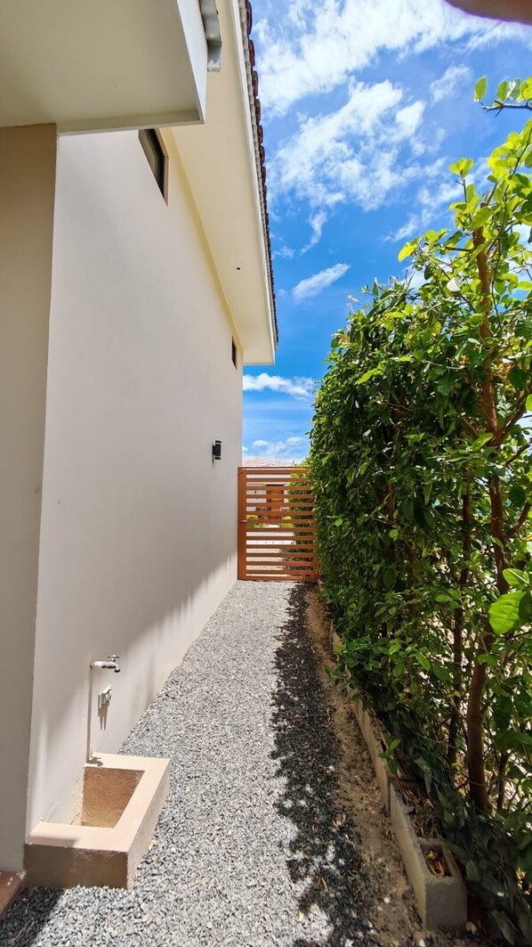 Ein schmaler Pfad führt zu einem Gebäude mit weißer Fassade, einem hölzernen Tor und einer Hecke.
