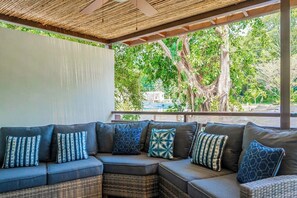 Ein überdachter Außenbereich mit Rattan-Sofa und gemusterten Kissen, mit Blick auf eine üppige Grünlandschaft.
