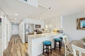 A modern kitchen with white cabinetry, a central island with stools, and a dining area with a white table. - Oceanfront condo with spacious private balcony, pool & direct beach access (North Myrtle Beach)
