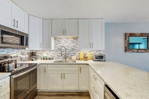 A modern kitchen with white cabinets, a tiled backsplash, stainless steel appliances, and a marble countertop. - Oceanfront condo with spacious private balcony, pool & direct beach access (North Myrtle Beach)