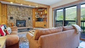 Ein gemütliches Wohnzimmer mit Kamin aus Stein, Holzregalen und einem großen Fenster mit Blick auf eine malerische Landschaft.