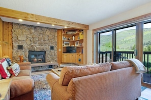 Ein gemütliches Wohnzimmer mit Kamin aus Stein, Holzregalen und einem großen Fenster mit Blick auf eine malerische Landschaft.