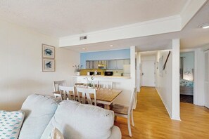 A modern living room with a sofa, dining table, and chairs, leading to a kitchen area with cabinets and a microwave. - Oceanview condo with pool, private balcony & direct beach access (Murrells Inlet)