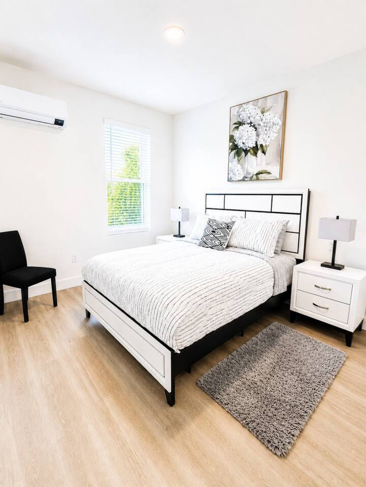 Un dormitorio con cama, mesitas de noche, una silla, un ventanal con persianas y aire acondicionado instalado en la pared.
