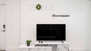 A modern living room with a flat-screen TV on a white marble cabinet, a small potted plant, and a 'HOME' sign with a wreath on the wall. - Stylish 1 BR Suite • Downtown • Convention Center (Hot Springs)