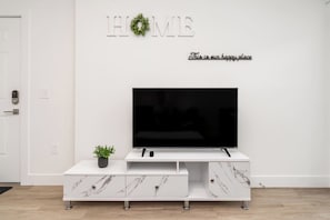 Ein modernes Wohnzimmer mit einem Flachbildfernseher auf einem weißen Marmorunterschrank, einer kleinen Topfpflanze und einem 'HOME'-Schild mit einem Kranz an der Wand.
