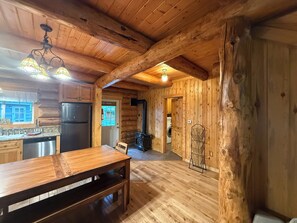 Interior de una cabaña con paredes de madera, una zona de cocina con refrigerador, una mesa de comedor y un banco.