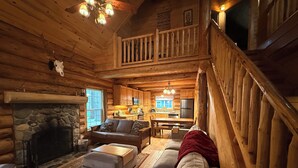 Interior de una cabaña de troncos con chimenea de piedra, muebles de madera, una zona de cocina y una escalera.