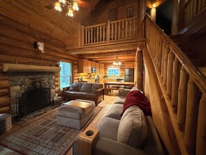 A log cabin interior with a stone fireplace, wooden furniture, a kitchen area, and a staircase.