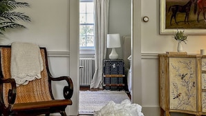 Une pièce comprenant une chaise en bois, une table blanche avec un vase, un meuble orné de motifs floraux et un tableau représentant des chevaux.
