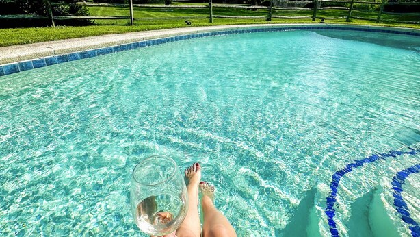 Une piscine aux eaux claires, avec un motif bleu sur le bord, une personne tenant un verre et, au fond, une zone herbeuse.