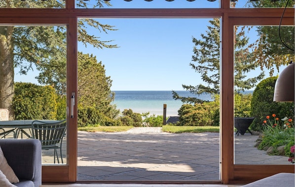 Vista a través de un ventanal a un patio con mesa y sillas, una terraza de madera y una playa al fondo.
