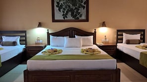 A hotel room with three beds, each with a wooden headboard, white bedding, and a green towel on the beds. There are two bedside lamps and a framed artwork on the wall.