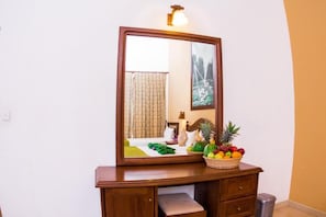 A wooden vanity with a mirror, a fruit basket, and a bed with a green bedspread and curtains.