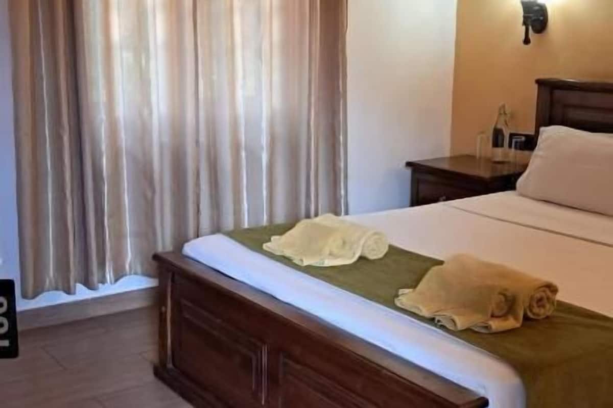 A hotel room with a wooden bed, a nightstand with a lamp, a window with curtains, and a wooden floor.
