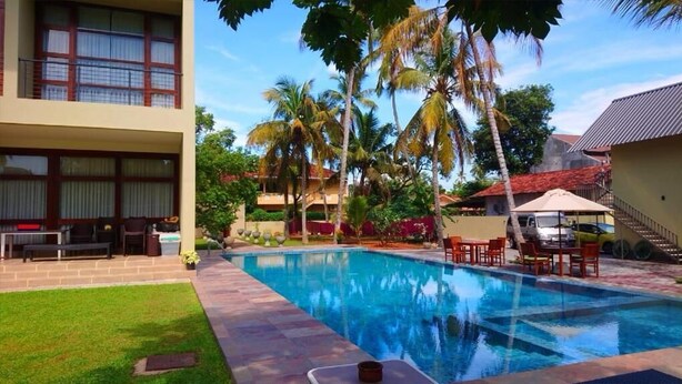 A swimming pool with lounge chairs and tables, surrounded by palm trees and a building with balconies. - jasmin villa ayurveda resort (Negombo)