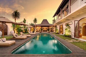 A pool area with lounge chairs and umbrellas, a dining area with tables and chairs, and a building with a thatched roof. - Villa Amara by Alfred in Bali (Seminyak)