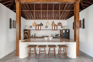 A modern kitchen with a central island, wooden stools, hanging pendant lights, and a tiled backsplash. - Villa Amara by Alfred in Bali (Seminyak)