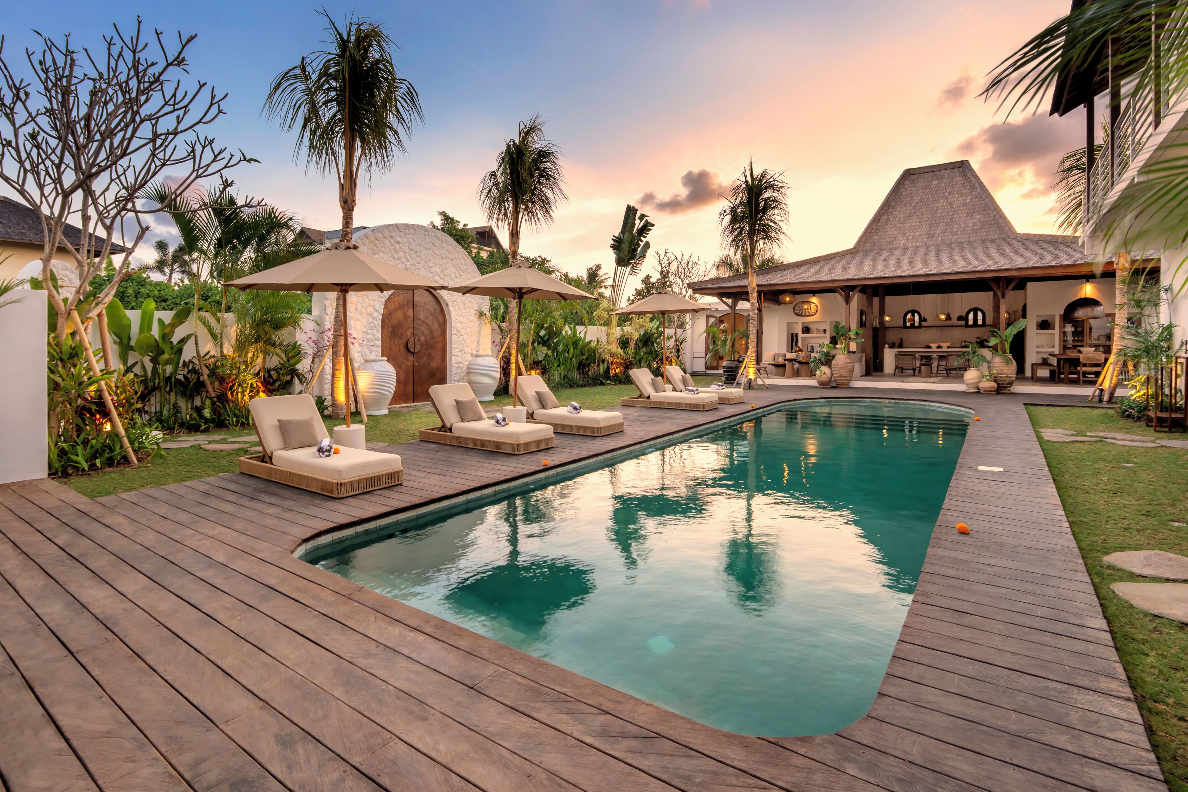 A pool area with lounge chairs and umbrellas, surrounded by tropical plants and a stone wall.