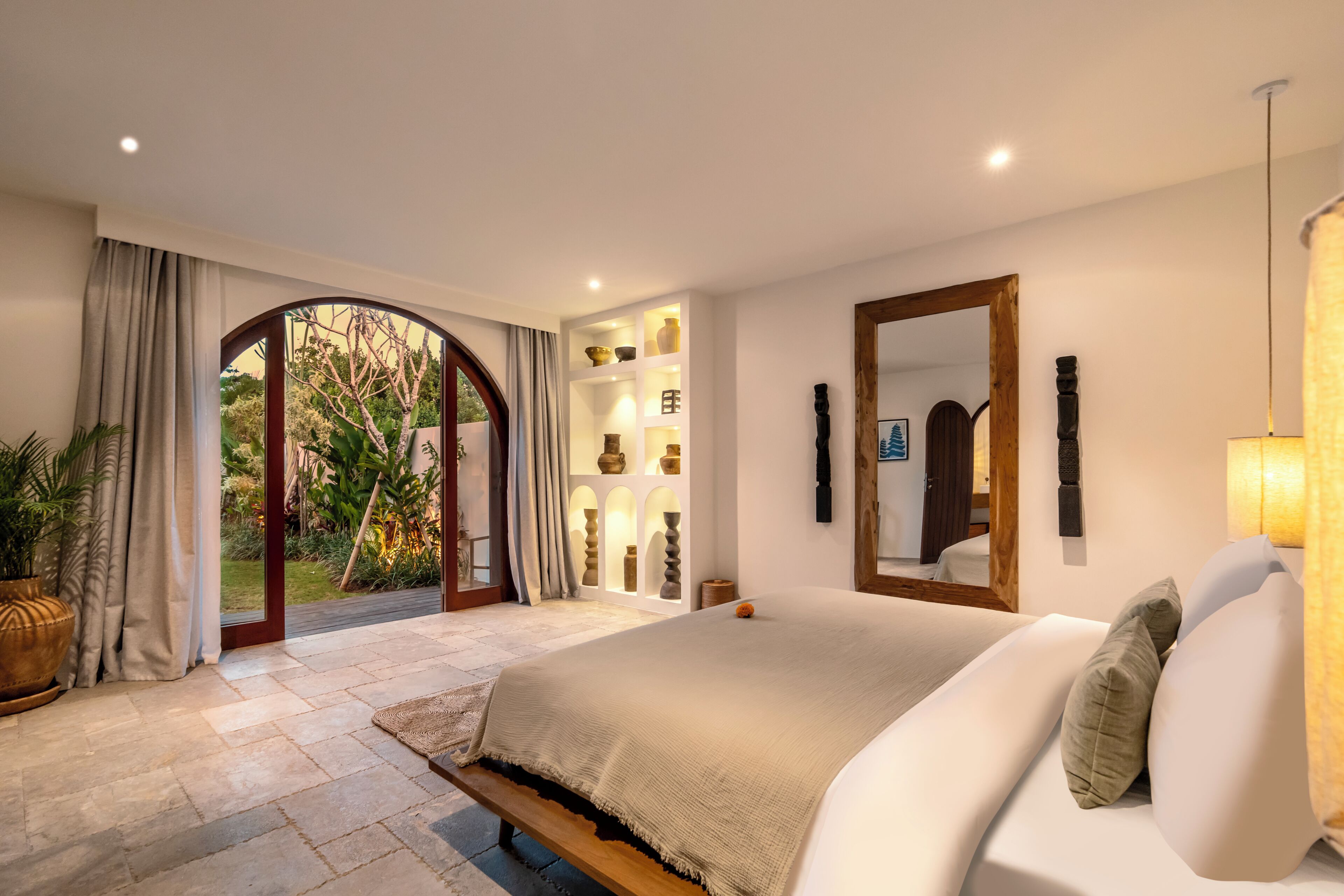 A bedroom with a large bed, a wooden bench, a built-in shelf with decorative items, and a view of a garden through an arched doorway.