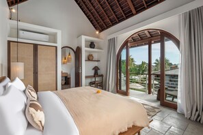 A bedroom with a large bed, a wardrobe, shelves, and a view of a garden through a glass door. - Villa Amara by Alfred in Bali (Seminyak)