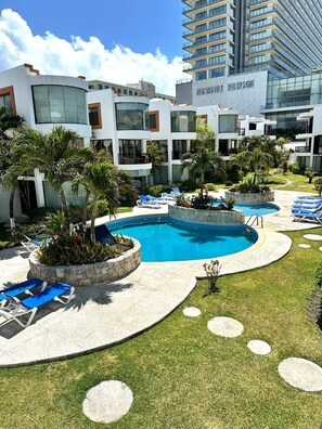 Zona de piscina de hotel con varias piscinas, sillones reclinables y palmeras.