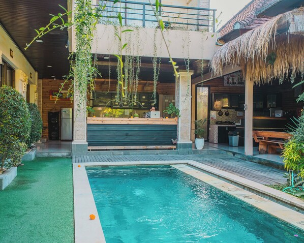 A pool area with a wooden deck, a bar area with a person working on a laptop, and a thatched roof structure.