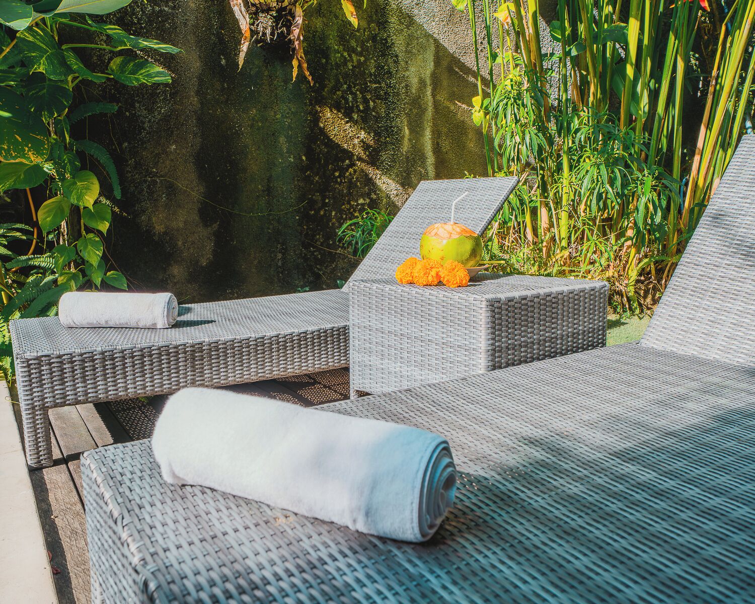A lounge area with wicker furniture, a rolled towel, a coconut drink, and a bowl of fruit.