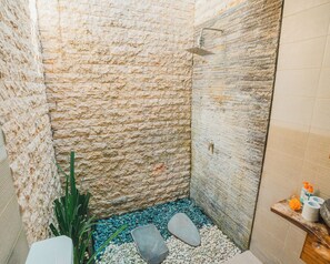 A stone-walled shower with a handheld showerhead, a small step leading into the shower, and a wooden shelf with toiletries.