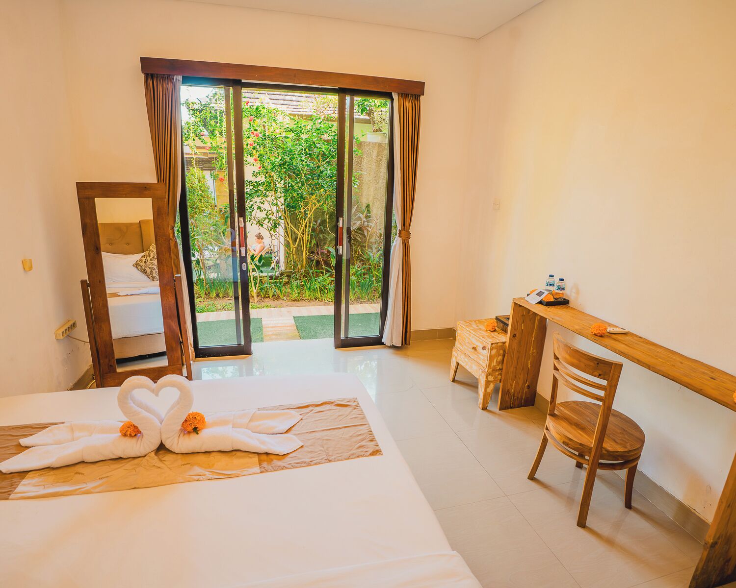 A hotel room with a bed, a desk, a chair, a bathroom, and a sliding glass door leading to a garden.
