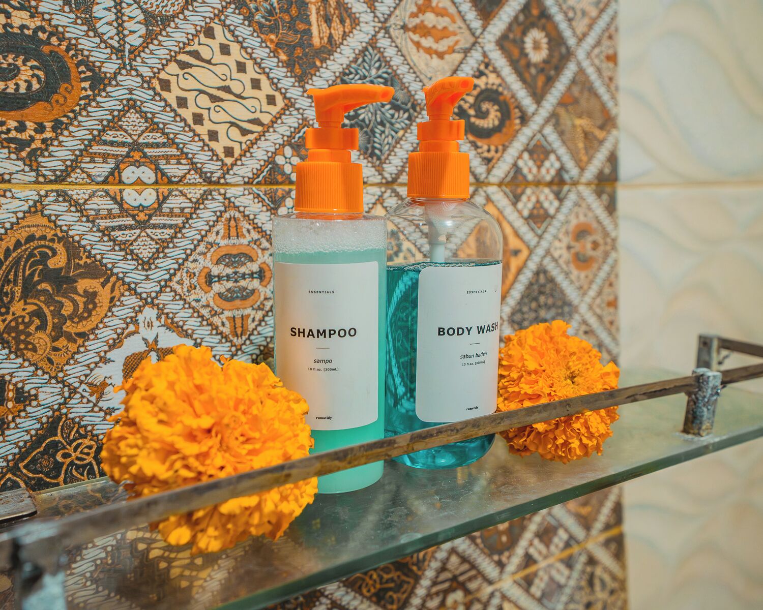 Deux bouteilles de shampoing et de gel douche sont posées sur une étagère en verre, avec des carreaux décoratifs et des fleurs de souci en arrière-plan.