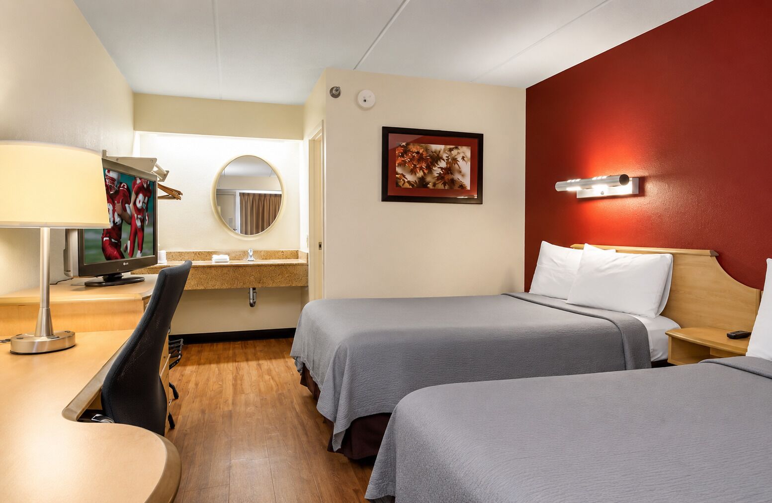 A hotel room with two beds, a desk with a TV, a mirror, and a red accent wall.