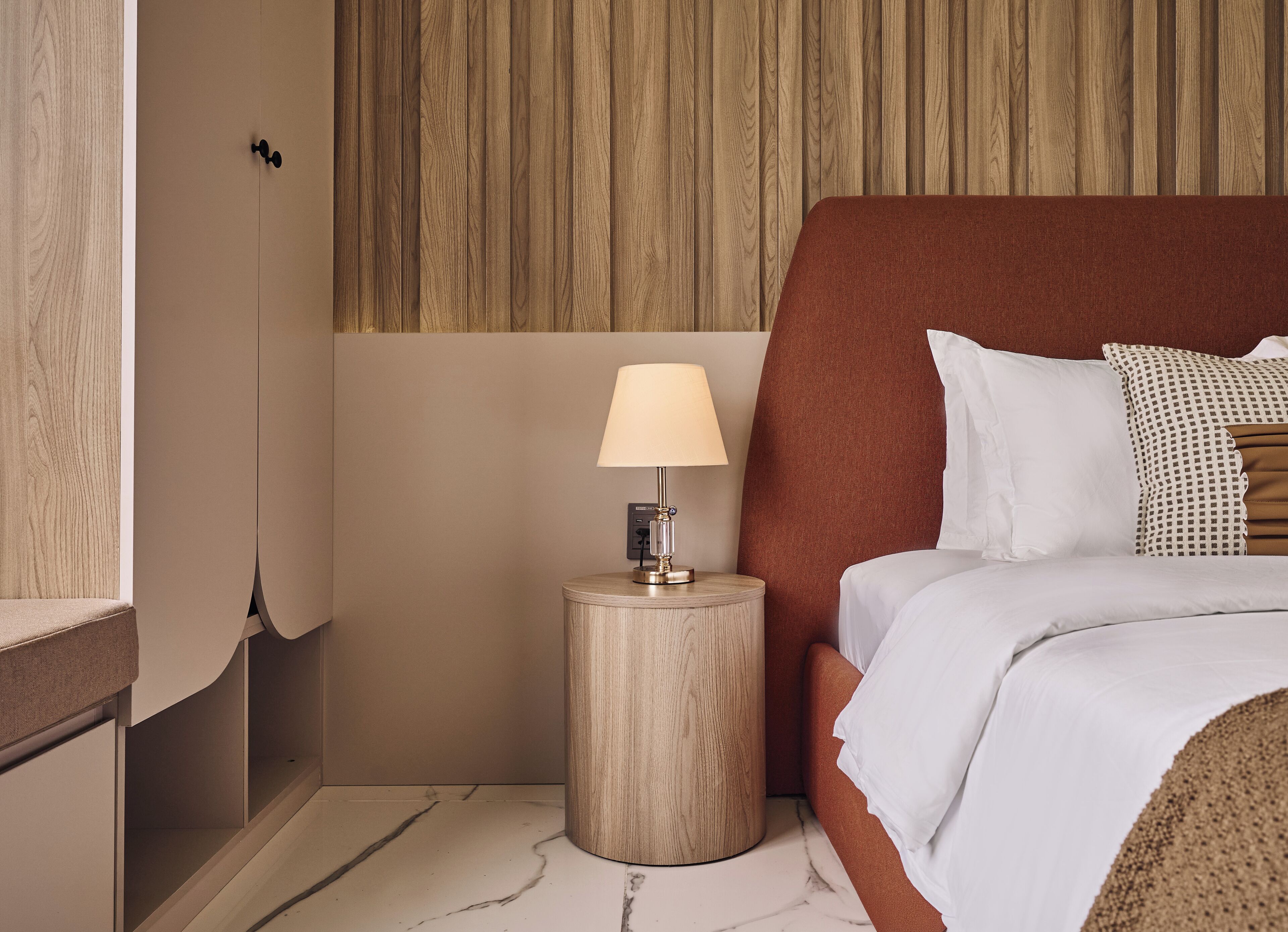 A hotel room with a bed, a nightstand with a lamp, a wooden headboard, and a marble floor.