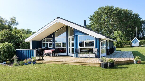 Ein modernes Haus mit großer Glasfront, einer überdachten Terrasse und einem kleinen Nebengebäude im Hintergrund.