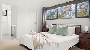 A bedroom with a bed, bedside tables, a window with curtains, and a door leading to another room. - Tea Tree Poolside Escape (Inverloch)