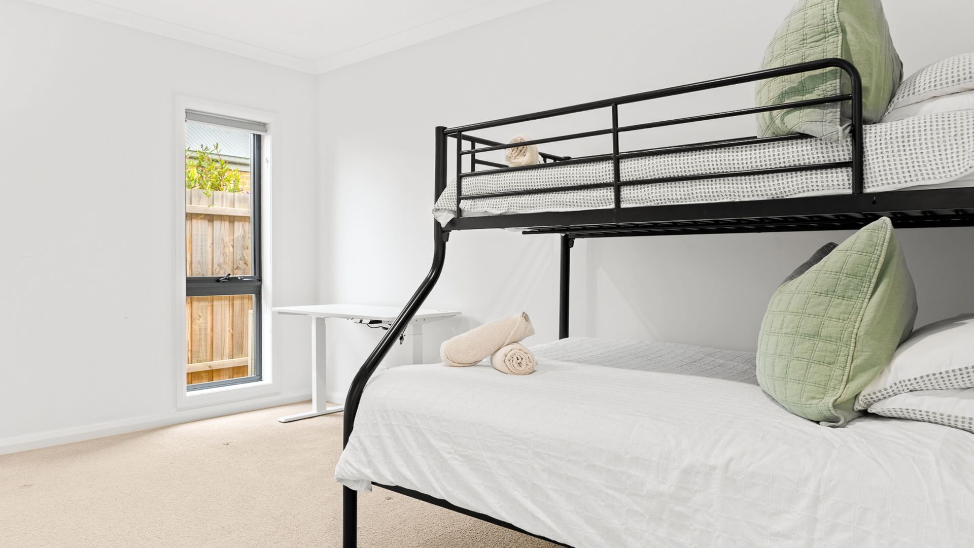 Ein Stockbett mit weißer Matratze und grünen Kissen, ein kleiner weißer Tisch und ein Fenster mit Blick auf einen Holenzaun.