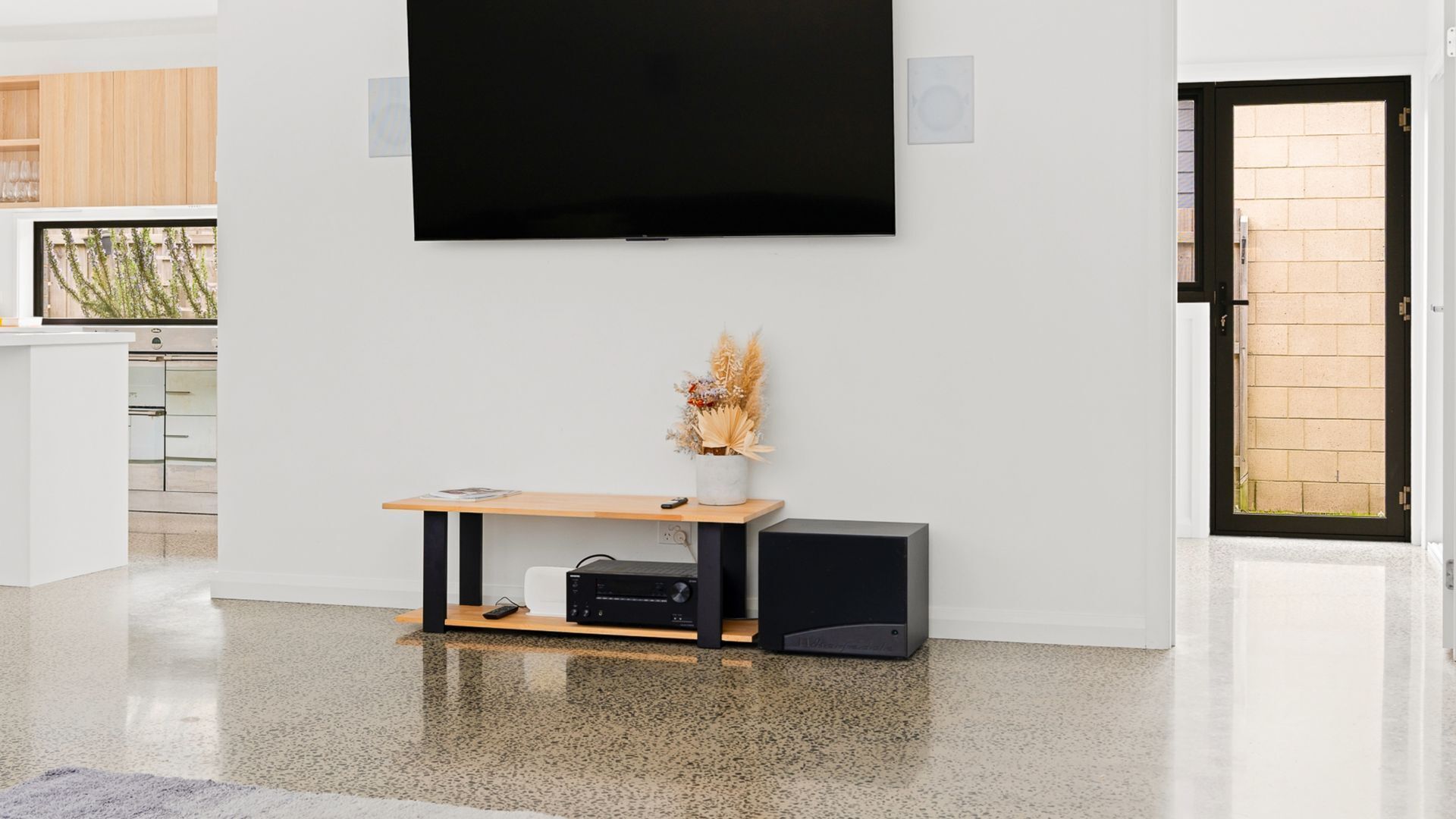 Een moderne woonkamer met een flatscreen televisie aan de muur, een houten salontafel met een vaas gedroogde planten, en een zwarte speaker.