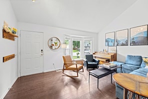 A living room with hardwood flooring, a rocking chair, a blue sofa, a piano, and framed pictures on the wall. - World Cup Home Base | 3BR + King Suite (Shawnee)