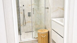 Een badkamer met een douche met glazen wand, een houten prullenbak en een wit wastafelmeubel.