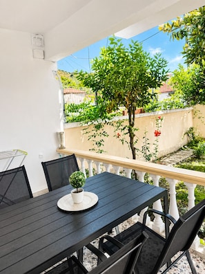 Um alpendre coberto com mesa e cadeiras, rodeado por vegetação e vista para uma área residencial.