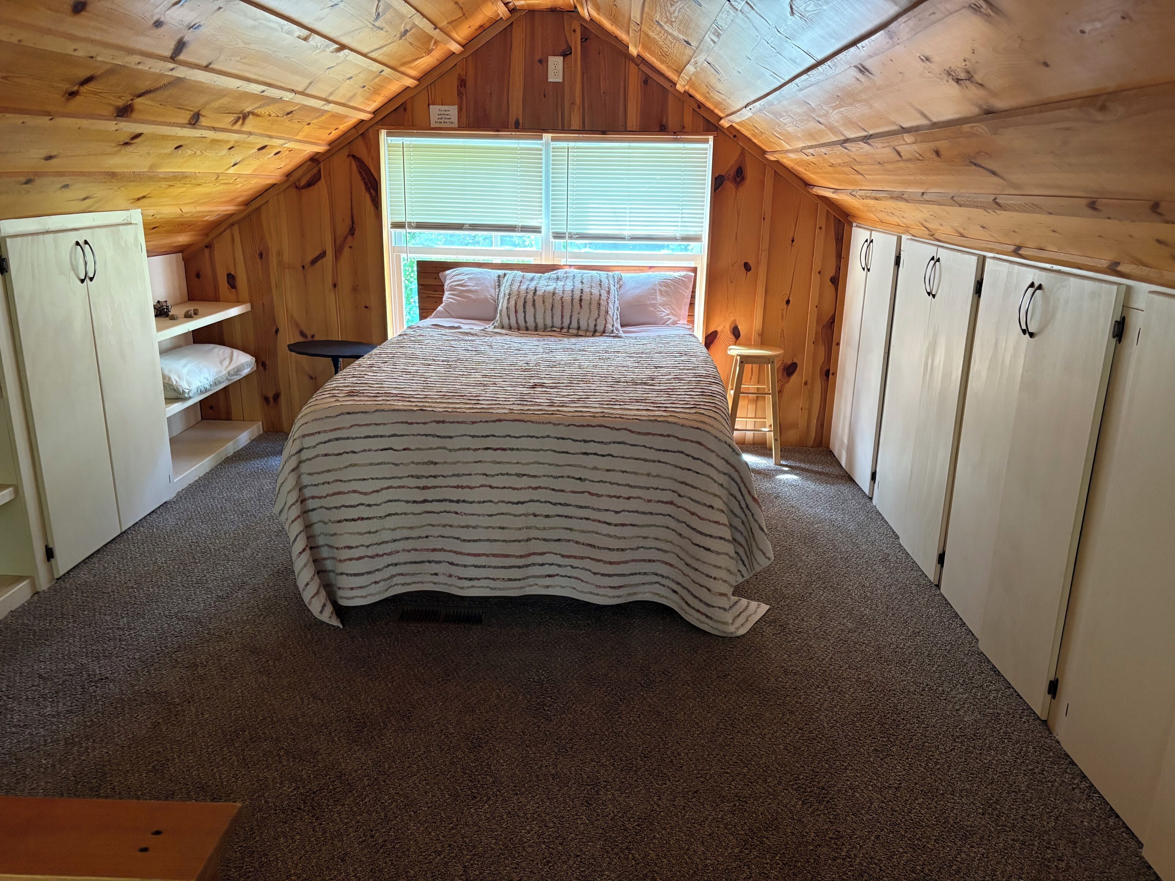 A bedroom with a sloped ceiling, a bed with striped bedding, a window with blinds, and wooden walls.
