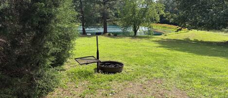 Un espacio cubierto de césped con una parrilla y una fogata, rodeado de árboles y una masa de agua.