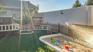 Zona de juegos al aire libre con cerramiento, arena, un banco y un pequeño árbol.