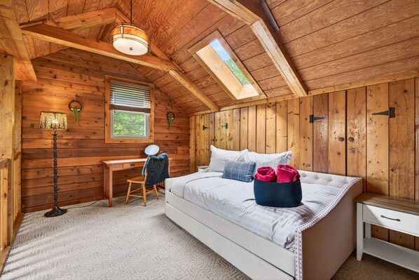 A cozy bedroom with a wooden ceiling, a desk, a chair, a bed with pillows, and a nightstand.