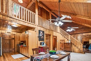 A wooden cabin interior with a dining area, kitchen, and staircase.