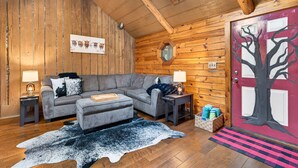 A cozy log cabin interior with a gray sectional sofa, a wooden ceiling, a red door with a tree mural, and a rustic wooden floor.