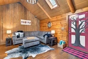 Ein gemütliches Blockhaus-Interieur mit einem grauen Ecksofa, einer Holzbalkendecke, einer roten Tür mit einem Baumgemälde und einem rustikalen Holzboden.